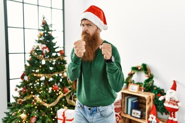 Redhead man with long beard wearing christmas hat by christmas tree ready to fight with fist defense gesture, angry and upset face, afraid of problem