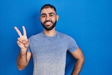 Middle east man with beard standing over blue background smiling looking to the camera showing fingers doing victory sign. number two.