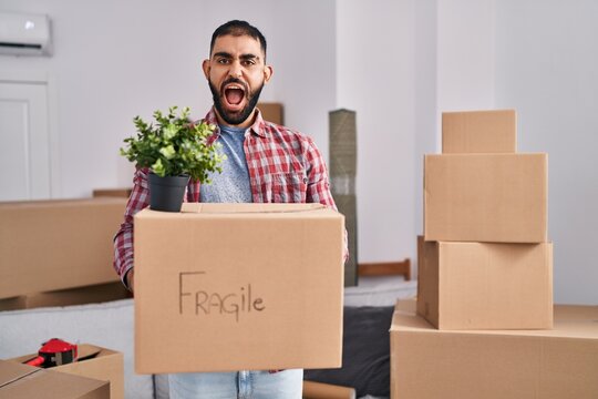 Middle east man with beard moving to a new home holding cardboard box angry and mad screaming frustrated and furious, shouting with anger. rage and aggressive concept.