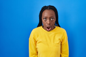 Beautiful black woman standing over blue background afraid and shocked with surprise and amazed...