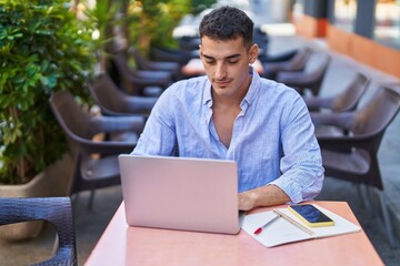 Young hispanic man using laptop sitting on table at coffee shop terrace