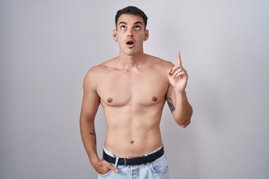 Handsome hispanic man standing shirtless amazed and surprised looking up and pointing with fingers and raised arms.