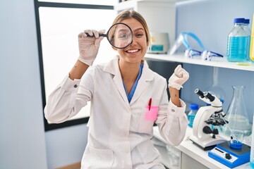 Young blonde woman working at scientist laboratory using magnifying glass screaming proud, celebrating victory and success very excited with raised arm
