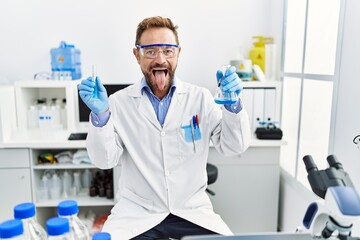 Middle age man working at scientist laboratory holding chemical products sticking tongue out happy...