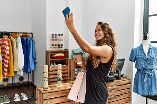 Young hispanic customer woman smiling happy holding shopping bags and make selfie by the smartphone at clothing store.