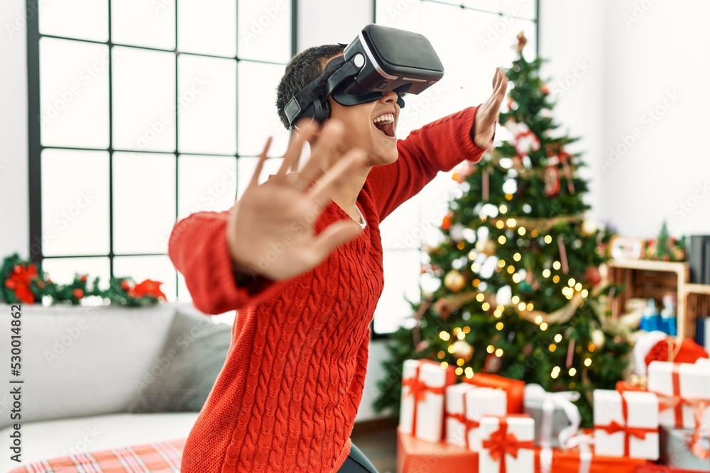 Sticker young hispanic woman playing video game using vr glasses standing by christmas tree at home