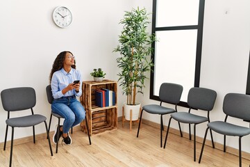 Young latin woman using smartphone sitting on chair at waiting room