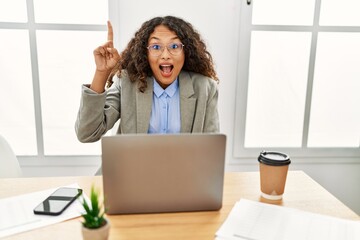 Beautiful hispanic business woman sitting on desk at office working with laptop pointing finger up with successful idea. exited and happy. number one.