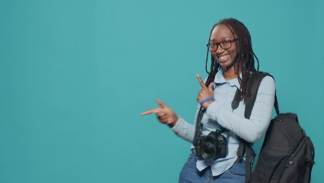 Photographer pointing at left and right side in studio, indicating sideways direction and carrying backpack with camera. Woman pointing lateral and aside, using photography equipment.