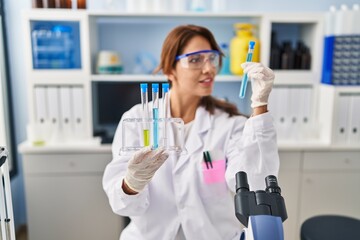 Young latin woman wearing scientist uniform holding test tubes at laboratory