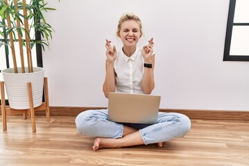 Young blonde woman using computer laptop sitting on the floor at the living room gesturing finger...