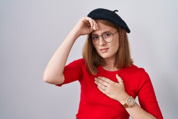 Young redhead woman standing wearing glasses and beret touching forehead for illness and fever, flu and cold, virus sick