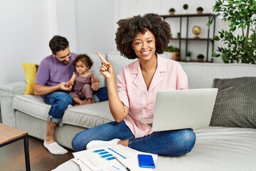 Mother of interracial family working using computer laptop at home with a big smile on face, pointing with hand finger to the side looking at the camera.