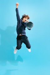 Portrait of young man with mustache posing, shouting in megaphone in a jump isolated over blue...