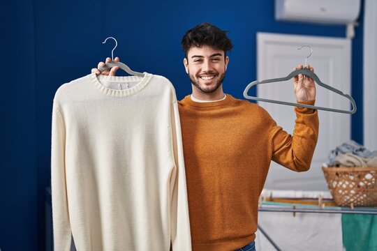 Hispanic man with beard holding sweater on hanger at laundry room smiling with a happy and cool smile on face. showing teeth.