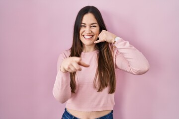 Young brunette woman standing over pink background smiling doing talking on the telephone gesture and pointing to you. call me.