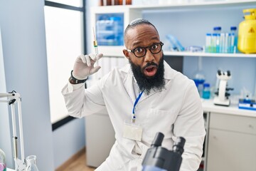 African american man working at scientist laboratory holding syringe scared and amazed with open mouth for surprise, disbelief face
