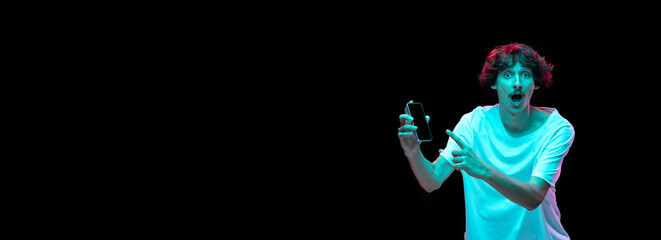 Portrait of young man with mustache pointing at phoen screen with emotions isolated over black background in neon light