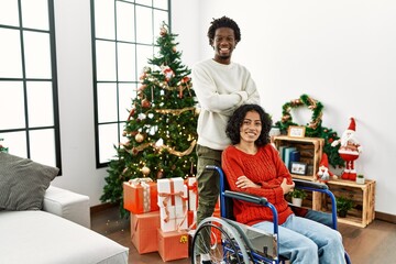 Young interracial couple with woman sitting on wheelchair by christmas tree happy face smiling with crossed arms looking at the camera. positive person.