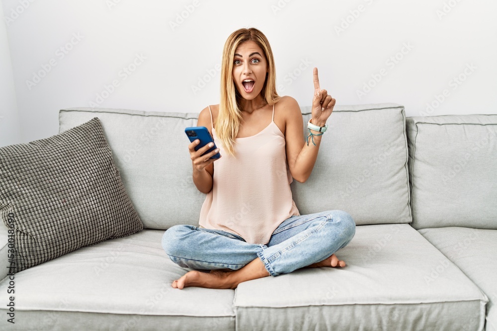 Canvas Prints Blonde beautiful young woman sitting on the sofa at home using smartphone pointing finger up with successful idea. exited and happy. number one.