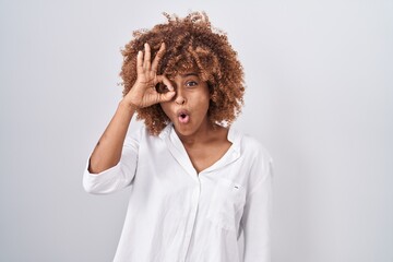 Young hispanic woman with curly hair standing over white background doing ok gesture shocked with surprised face, eye looking through fingers. unbelieving expression.