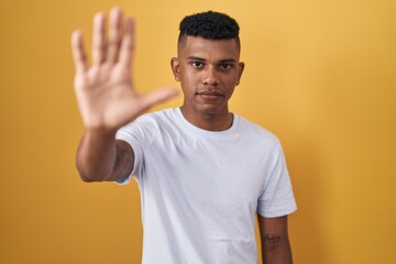 Young hispanic man standing over yellow background doing stop sing with palm of the hand. warning expression with negative and serious gesture on the face.