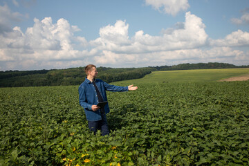 A farmer with a digital tablet checks the quality of soybeans in an agricultural field and points with his hand into the distance. Front view