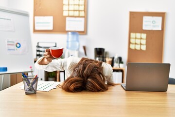 Young hispanic businesswoman overworked with head on table and holding cup of coffee at the office.