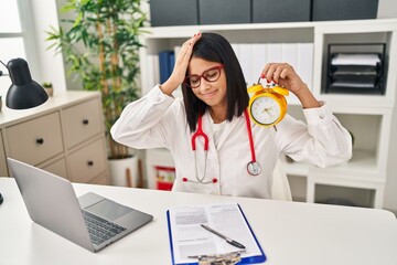 Young hispanic doctor woman holding alarm clock at the clinic stressed and frustrated with hand on head, surprised and angry face