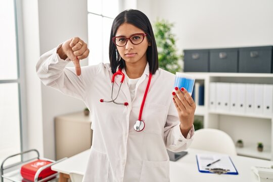 Young hispanic doctor woman holding cotton buds with angry face, negative sign showing dislike with thumbs down, rejection concept