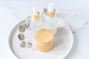 Jar of organic cream, face serum in dorpper bottle with eucalyptus leaves on a tray on marble. Unbranded mockup cosmetics set. Natural organic product, beauty and spa concept