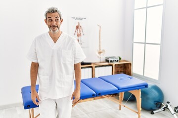 Middle age hispanic therapist man working at pain recovery clinic smiling looking to the side and staring away thinking.