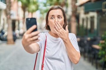 Middle age hispanic woman doing video call with smartphone covering mouth with hand, shocked and...