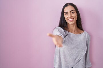 Fototapeta premium Young brunette woman standing over pink background smiling friendly offering handshake as greeting and welcoming. successful business.