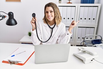 Young hispanic woman wearing doctor uniform holding stethoscope at medical clinic smiling happy pointing with hand and finger to the side