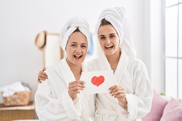 Middle age woman and daughter wearing bath robe holding heart card smiling and laughing hard out...