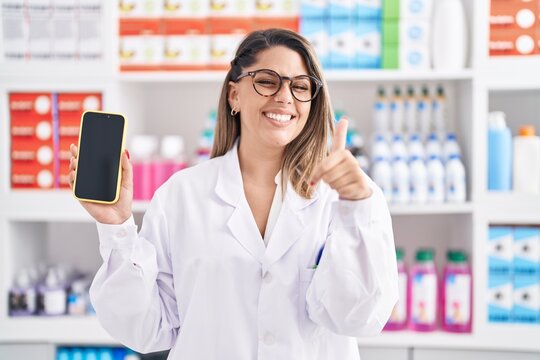 Blonde woman working at pharmacy drugstore showing smartphone screen smiling happy and positive, thumb up doing excellent and approval sign