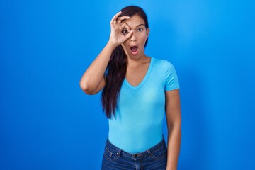 Young hispanic woman standing over blue background doing ok gesture shocked with surprised face, eye looking through fingers. unbelieving expression.