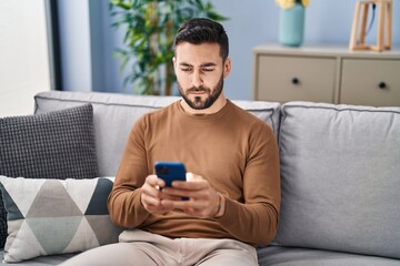 Young hispanic man using smartphone sitting on sofa at home