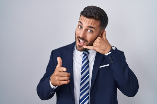 Handsome hispanic man wearing suit and tie smiling doing talking on the telephone gesture and pointing to you. call me.