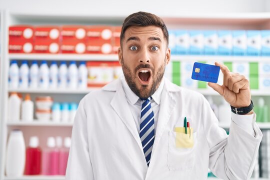 Handsome hispanic man working at pharmacy drugstore holding credit card scared and amazed with open mouth for surprise, disbelief face