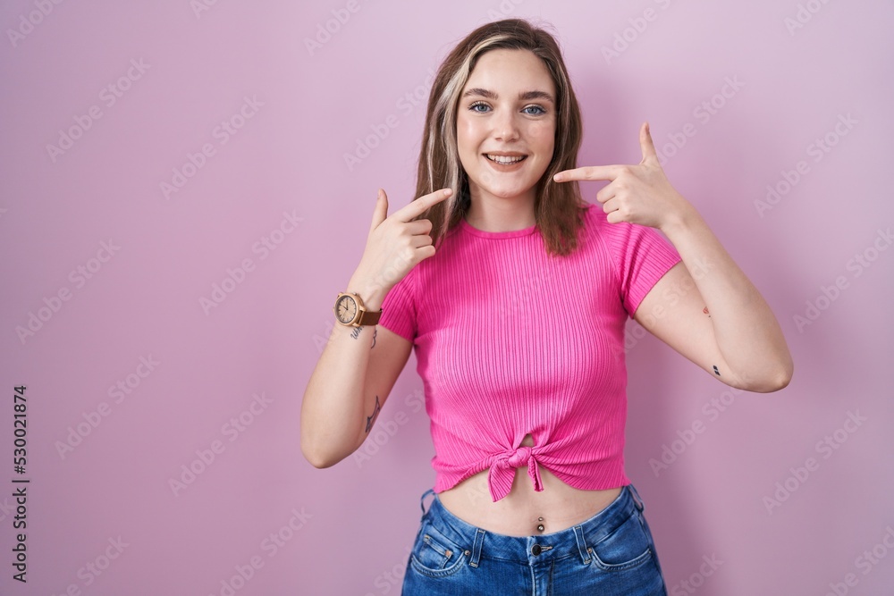 Sticker Blonde caucasian woman standing over pink background smiling cheerful showing and pointing with fingers teeth and mouth. dental health concept.