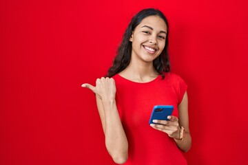 Naklejka premium Young brazilian woman using smartphone over red background pointing to the back behind with hand and thumbs up, smiling confident