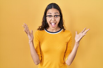 Young brazilian woman wearing glasses over yellow background celebrating victory with happy smile and winner expression with raised hands