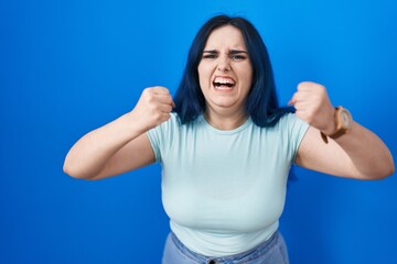 Young modern girl with blue hair standing over blue background angry and mad raising fists frustrated and furious while shouting with anger. rage and aggressive concept.