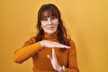 Middle age hispanic woman standing over yellow background doing time out gesture with hands,...