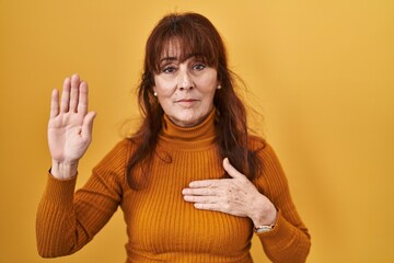 Middle age hispanic woman standing over yellow background swearing with hand on chest and open...