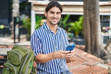 Young hispanic man tourist listening to music sitting on bench at park