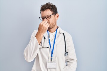 Young hispanic man wearing doctor uniform and stethoscope smelling something stinky and disgusting,...