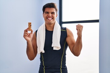 Hispanic man eating protein bar as healthy energy snack screaming proud, celebrating victory and...